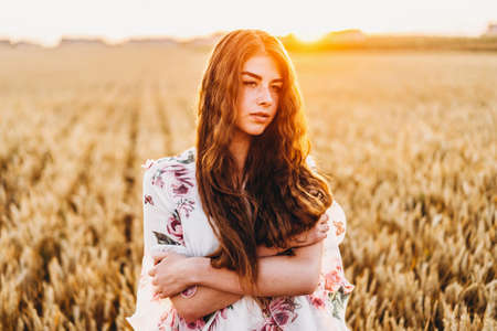 Portrait of a beautiful young woman with curly hair and freckles face. Woman in dress and hat posing in wheat field at sunset and looking at camera.の写真素材