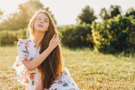 Portrait of a beautiful young woman with curly hair and freckles face. Woman in dress and hat posing in wheat field at sunset and looking at camera.の写真素材