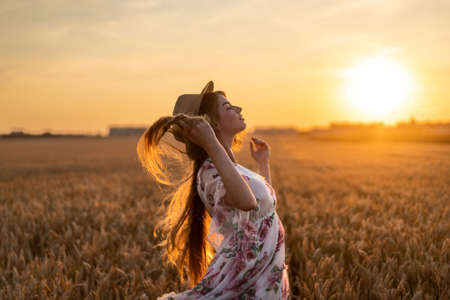 Portrait of a beautiful young woman with curly hair and freckles face. Woman in dress and hat posing in wheat field at sunset and looking at camera.の写真素材