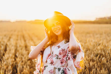 Portrait of a beautiful young woman with curly hair and freckles face. Woman in dress and hat posing in wheat field at sunset and looking at camera.の写真素材