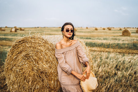 Brunette in linen pants and bare shoulders sitting on a hay bales in warm autumn day. Woman looking...