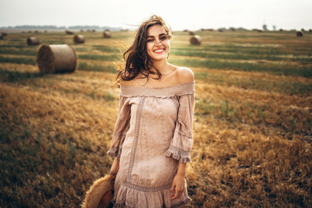Brunette in linen pants and bare shoulders sitting on a hay bales in warm autumn day. Woman looking at camera. Behind her is a wheat field.の写真素材