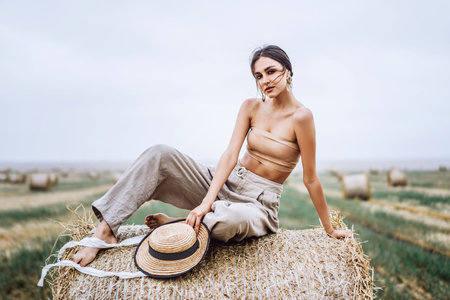 Brunette in linen pants and bare shoulders sitting on a hay bales in warm autumn day. Woman looking at camera. Behind her is a wheat field.の写真素材