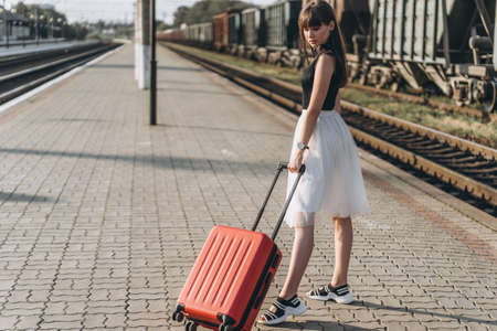 Female brunette traveler with red suitcase in white skirt waiting for a train and walking on raiway stationの写真素材