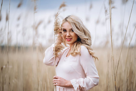 Photo of a pretty smiling girl with long blond curly hair in light long drees standing in a reed field and holding high reed branches in her handsの写真素材
