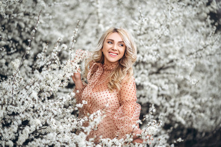 Photo of a pretty smiling girl with long blond curly hair in light long drees standing in a reed field and holding high reed branches in her handsの写真素材