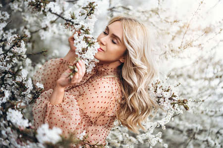 Photo of a pretty smiling girl with long blond curly hair in light long drees standing in a reed field and holding high reed branches in her handsの写真素材