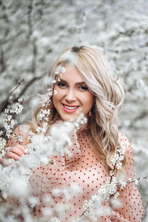 Photo of a pretty smiling girl with long blond curly hair in light long drees standing in a reed field and holding high reed branches in her handsの写真素材