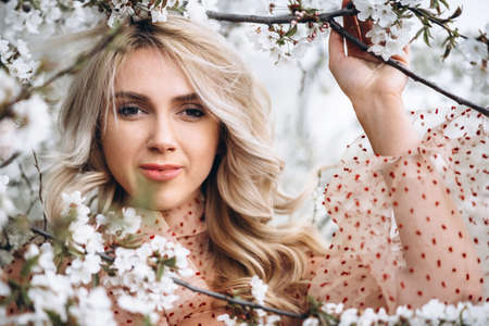 Photo of a pretty smiling girl with long blond curly hair in light long drees standing in a reed field and holding high reed branches in her handsの写真素材