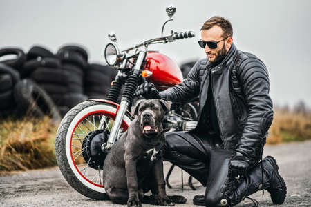 Bearded man in sunglasses and leather jacket looking at the camera while sitting on a motorcycle on the road. Behind him is a row of tires.の写真素材