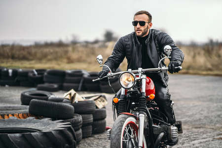 Bearded man in sunglasses and leather jacket looking at the camera while sitting on a motorcycle on the road. Behind him is a row of tires.の写真素材