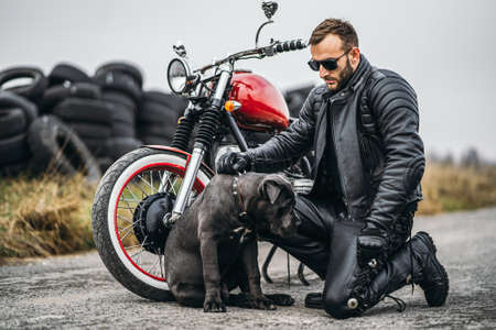 Bearded man in sunglasses and leather jacket looking at the camera while sitting on a motorcycle on the road. Behind him is a row of tires.の写真素材
