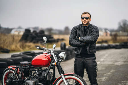 Bearded man in sunglasses and leather jacket looking at the camera while sitting on a motorcycle on the road. Behind him is a row of tires.の写真素材
