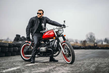 Bearded man in sunglasses and leather jacket looking at the camera while sitting on a motorcycle on the road. Behind him is a row of tires.の写真素材