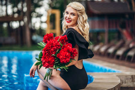 Pretty pin up styled blonde in black vintage swimsuit and hairstyle, with red lips, holding umbrella and relaxing in the swimming poolの写真素材