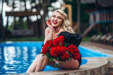 Pretty pin up styled blonde in black vintage swimsuit and hairstyle, with red lips, holding umbrella and relaxing in the swimming poolの写真素材