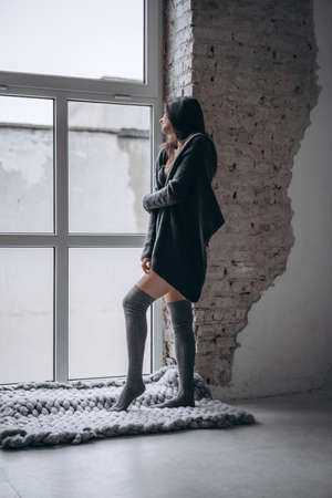Young brunette in black underwear, knitted knee pads and cardigan standing near window and brick wall in a warm blanket.の写真素材