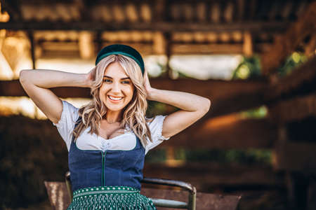 Pretty happy blonde in dirndl, traditional festival dress, holding two mugs of beer outdoors in the forest with blurred background. Oktoberfest, St. Patrickâs day, international beer day concept.の写真素材