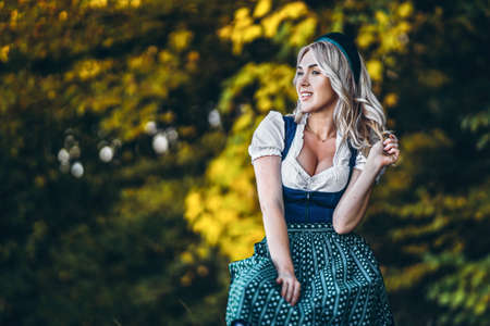 Pretty happy blonde in dirndl, traditional festival dress, holding two mugs of beer outdoors in the forest with blurred background. Oktoberfest, St. Patrickâs day, international beer day concept.の写真素材