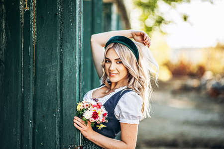 Pretty happy blonde in dirndl, traditional festival dress, holding two mugs of beer outdoors in the forest with blurred background. Oktoberfest, St. Patrickâs day, international beer day concept.の写真素材