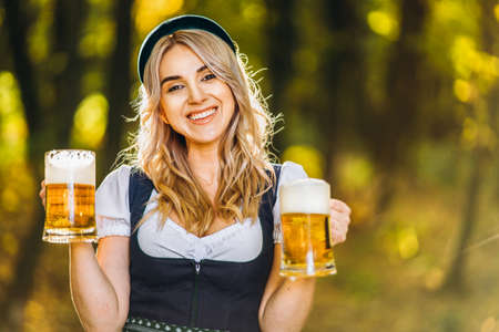 Pretty happy blonde in dirndl, traditional festival dress, holding two mugs of beer outdoors in the forest with blurred background. Oktoberfest, St. Patrickâs day, international beer day concept.の写真素材