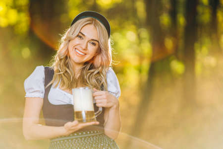 Pretty happy blonde in dirndl, traditional festival dress, holding two mugs of beer outdoors in the forest with blurred background. Oktoberfest, St. Patrickâs day, international beer day concept.の写真素材