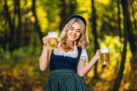 Pretty happy blonde in dirndl, traditional festival dress, holding two mugs of beer outdoors in the forest with blurred background. Oktoberfest, St. Patrickâs day, international beer day concept.の写真素材
