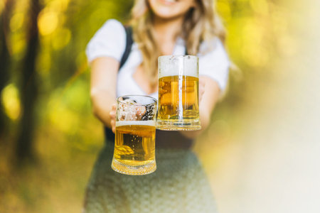 Pretty happy blonde in dirndl, traditional festival dress, holding two mugs of beer outdoors in the forest with blurred background. Oktoberfest, St. Patrickâs day, international beer day concept.の写真素材