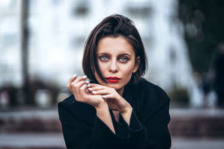 Close up portrait of young woman with short haircut and red lips, dressed in black. Woman posing, leaning on a wooden fence with plants. Blurred urban background.の写真素材