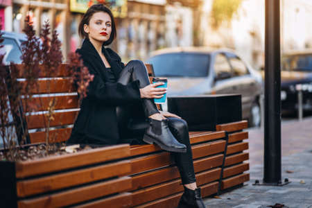 Close up portrait of young woman with short haircut and red lips, dressed in black. Woman posing, leaning on a wooden fence with plants. Blurred urban background.の写真素材