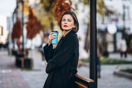 Close up portrait of young woman with short haircut and red lips, dressed in black. Woman posing, leaning on a wooden fence with plants. Blurred urban background.の写真素材