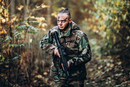 Portrait of a camouflage soldier with rifle and painted face playing airsoft outdoors in the forest. Side view.の写真素材