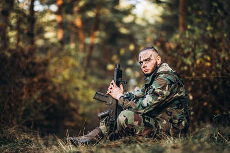 Portrait of a camouflage soldier with rifle and painted face playing airsoft outdoors in the forest. Side view.の写真素材