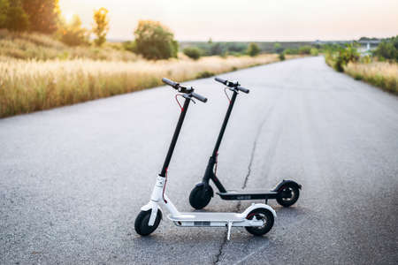 Two electric scooters, black and white, stand in the middle of the road in the countryside. Content technologies. Sunset time. New movement.の写真素材