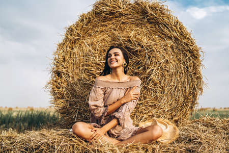 Closeup portrait of beautiful smiling woman with closed eyes. The brunette leaned on a bale of hay. A wheat field on the background.の写真素材
