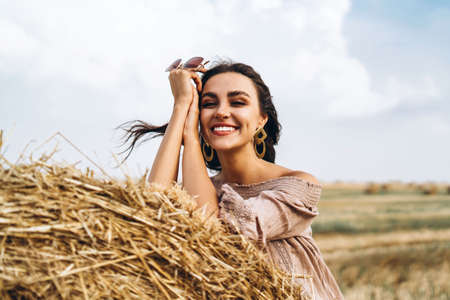 Closeup portrait of beautiful smiling woman with closed eyes. The brunette leaned on a bale of hay. A wheat field on the background.の写真素材