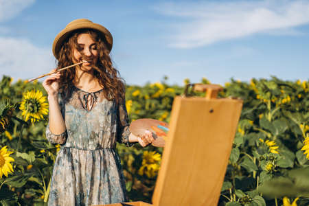 Closeup portrait of a beautiful young woman with curly hair. Woman in dress and hat standing in wheat field.の写真素材
