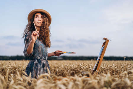 Closeup portrait of a beautiful young woman with curly hair. Woman in dress and hat standing in wheat field.の写真素材