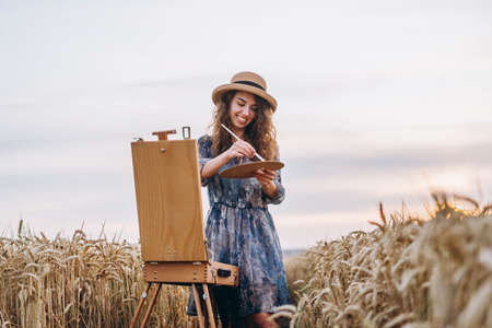 Closeup portrait of a beautiful young woman with curly hair. Woman in dress and hat standing in wheat field.の写真素材