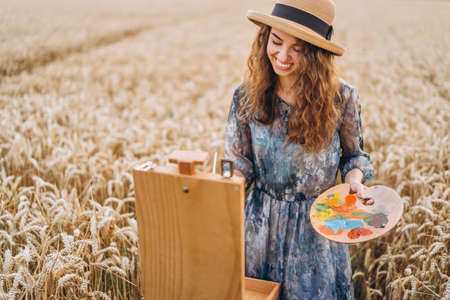 Closeup portrait of a beautiful young woman with curly hair. Woman in dress and hat standing in wheat field.の写真素材