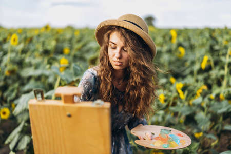 Closeup portrait of a beautiful young woman with curly hair. Woman in dress and hat standing in wheat field.の写真素材