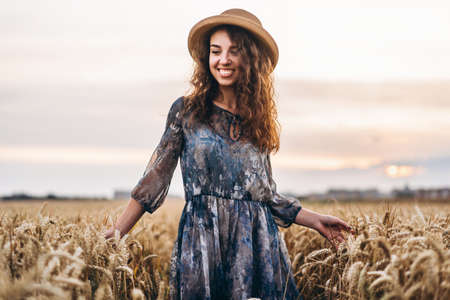 Closeup portrait of a beautiful young woman with curly hair. Woman in dress and hat standing in wheat field.の写真素材