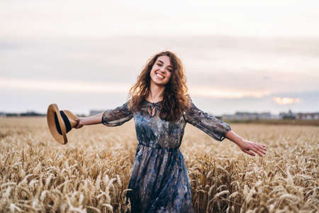 Closeup portrait of a beautiful young woman with curly hair. Woman in dress and hat standing in wheat field.の写真素材