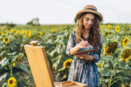 Closeup portrait of a beautiful young woman with curly hair. Woman in dress and hat standing in wheat field.の写真素材
