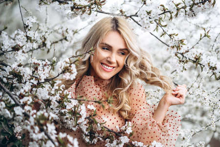 Photo of a pretty smiling girl with long blond curly hair in light long drees standing in a reed field and holding high reed branches in her handsの写真素材
