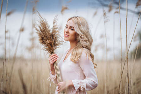 Photo of a pretty smiling girl with long blond curly hair in light long drees standing in a reed field and holding high reed branches in her handsの写真素材