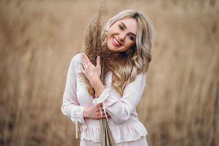 Photo of a pretty smiling girl with long blond curly hair in light long drees standing in a reed field and holding high reed branches in her handsの写真素材