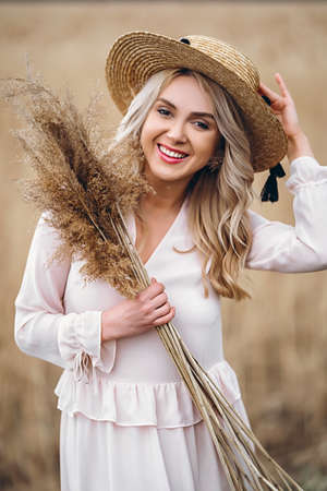 Photo of a pretty smiling girl with long blond curly hair in light long drees standing in a reed field and holding high reed branches in her handsの写真素材