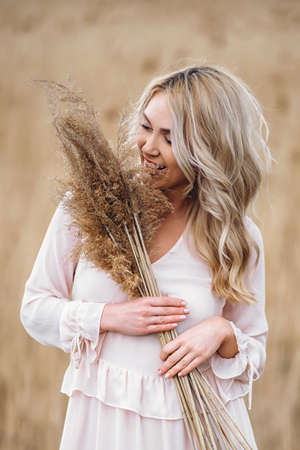 Photo of a pretty smiling girl with long blond curly hair in light long drees standing in a reed field and holding high reed branches in her handsの写真素材