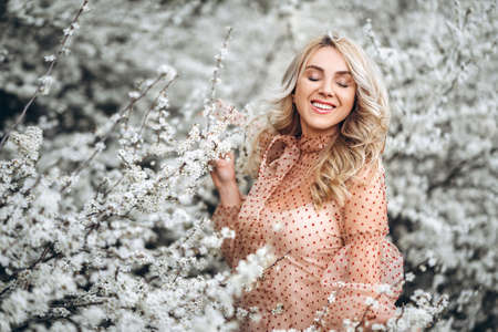 Photo of a pretty smiling girl with long blond curly hair in light long drees standing in a reed field and holding high reed branches in her handsの写真素材